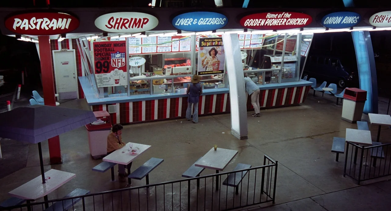 Straight Time (1978) movie still: wide — An outdoor fast food stand at night with red and blue neon signs. Patrons are eating at picnic tables and…