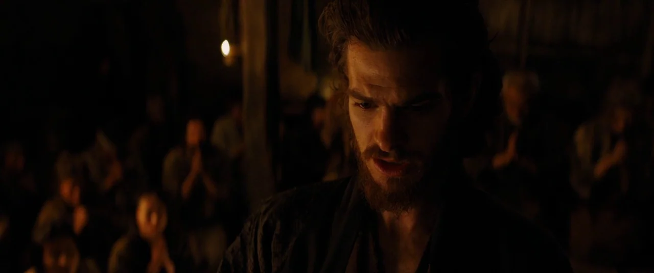 Silence (2016) movie still: closeup — A dark, cramped interior space filled with blurred figures in prayer. A man with a beard looks down in…