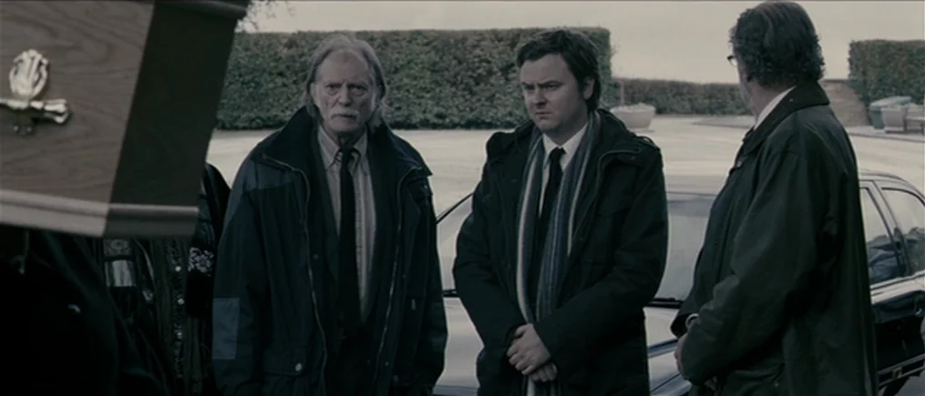Another Year (2010) movie still: medium — A paved area outside a graveyard features a polished black vehicle and manicured hedges. Three men stand…