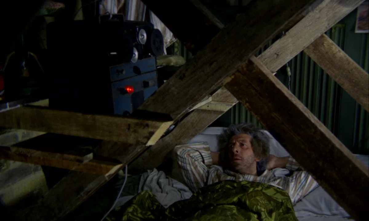 Wonderwall (1968) movie still: medium — A cluttered attic space features exposed wooden rafters positioned over a bed. A middle-aged man lies in bed…