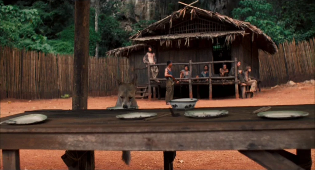 Rescue Dawn movie still: wide — A rustic settlement features a wooden hut with a thatched roof in a jungle clearing. A group of men stands or sits…