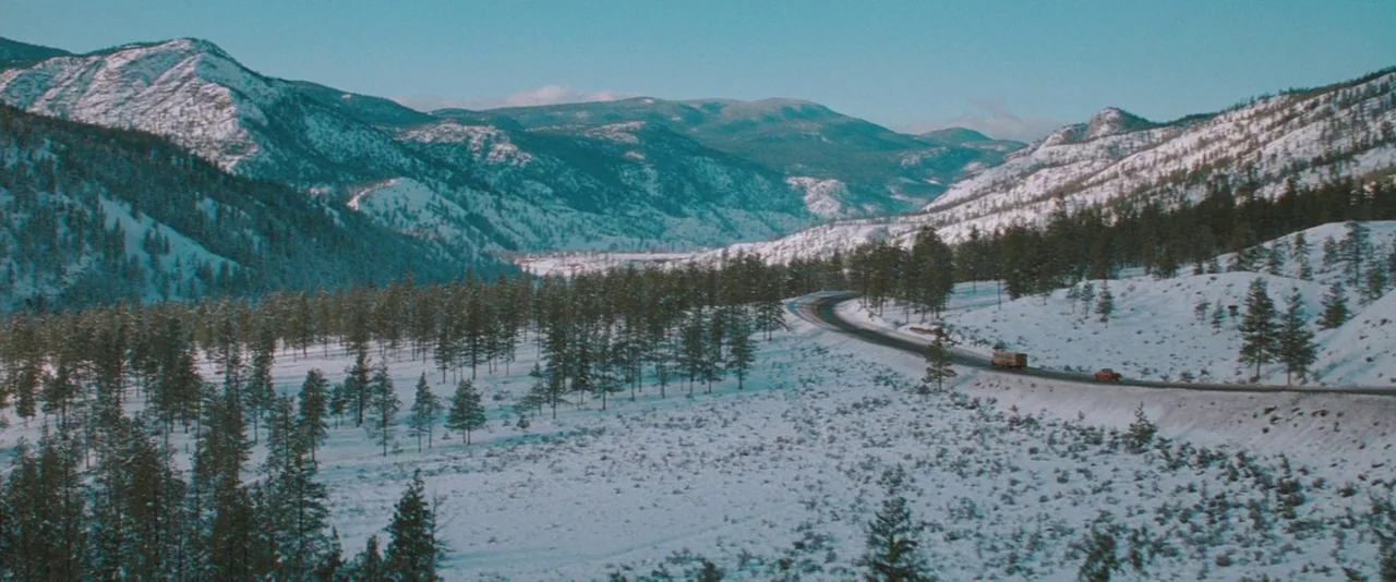 The Sweet Hereafter (1997) movie still: establishing — A vast, snowy valley is surrounded by pine-covered mountains under a clear sky. A narrow road…