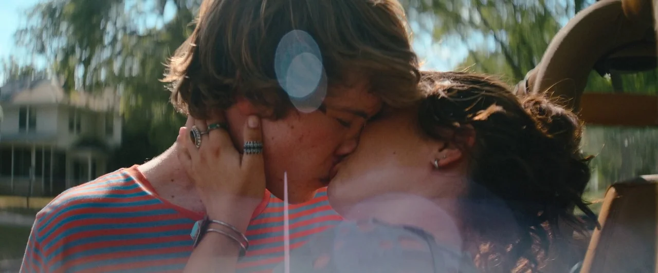 Snack Shack (2024) movie still: closeup — A man and a woman kiss in front of a residential house with green trees. The couple is shown in a tight…
