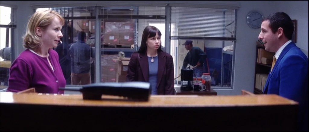 Punch Drunk Love (2002) movie still: medium — A brightly lit business front desk features a wooden counter and large glass windows. Two women stand…