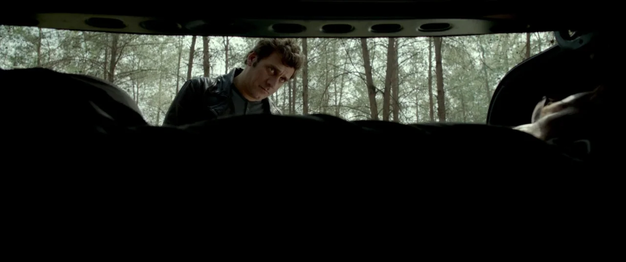 Big Bad Wolves (2013) movie still: medium — A man peers into the dark open trunk of a car parked in a forest. The perspective is from inside the…