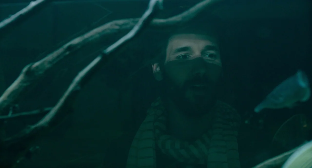 Confess, Fletch (2022) movie still: closeup — A man looks through the glass of a dark, green-lit aquatic display. A submerged wooden branch sits in…