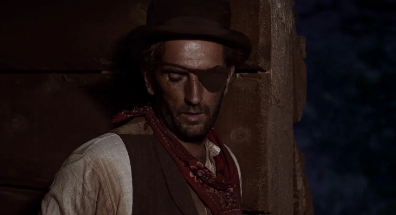 Ride In The Whirlwind (1966) movie still: medium — A man stands inside a dimly lit structure made of rough-hewn horizontal timber logs. The subject…