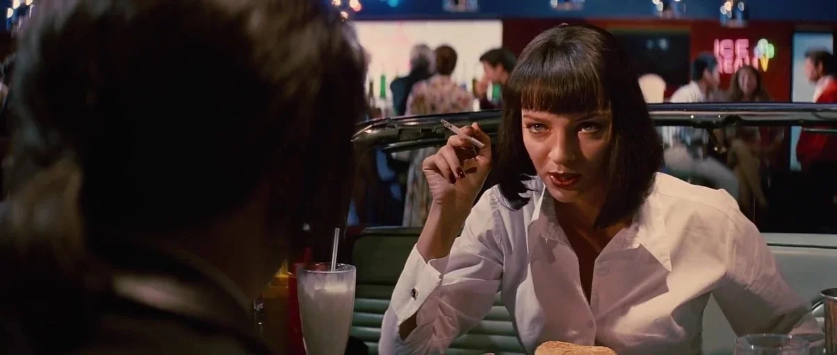 Pulp Fiction (1994) movie still: medium — A retro-themed 1950s style diner with green vinyl booths and overhead hanging lights. A woman sits in a…