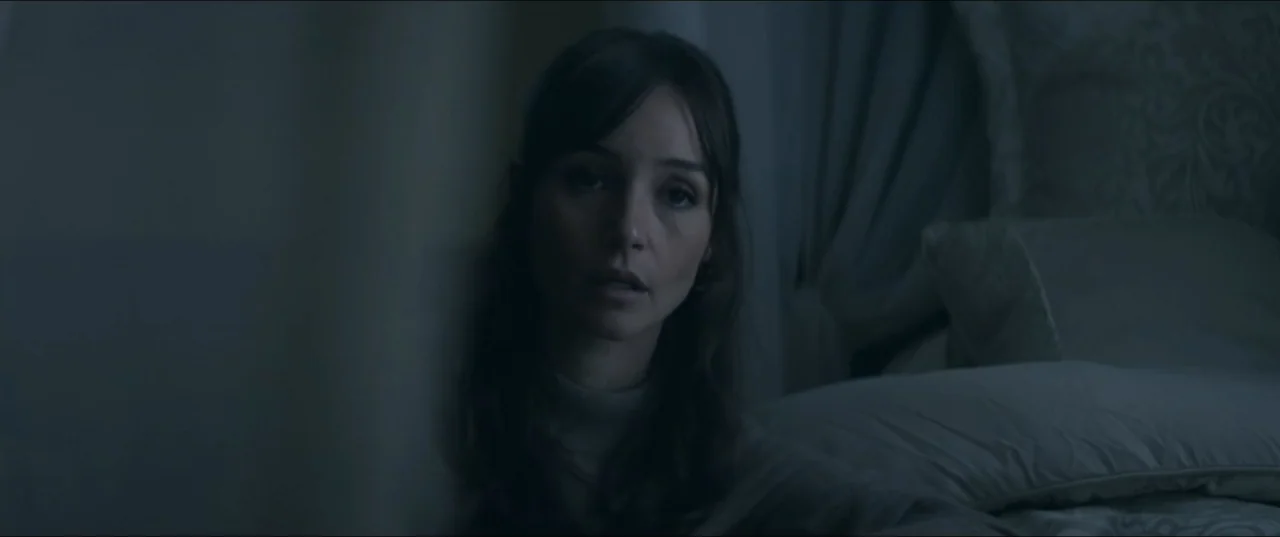Offseason (2022) movie still: closeup — A dark, dimly lit indoor space features a person positioned next to bedding. A woman is partially visible in…