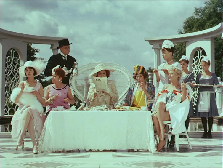 All These Women (1964) movie still: wide — An ornate white marble patio features an elegant outdoor tea setting under clouds. Several people in…