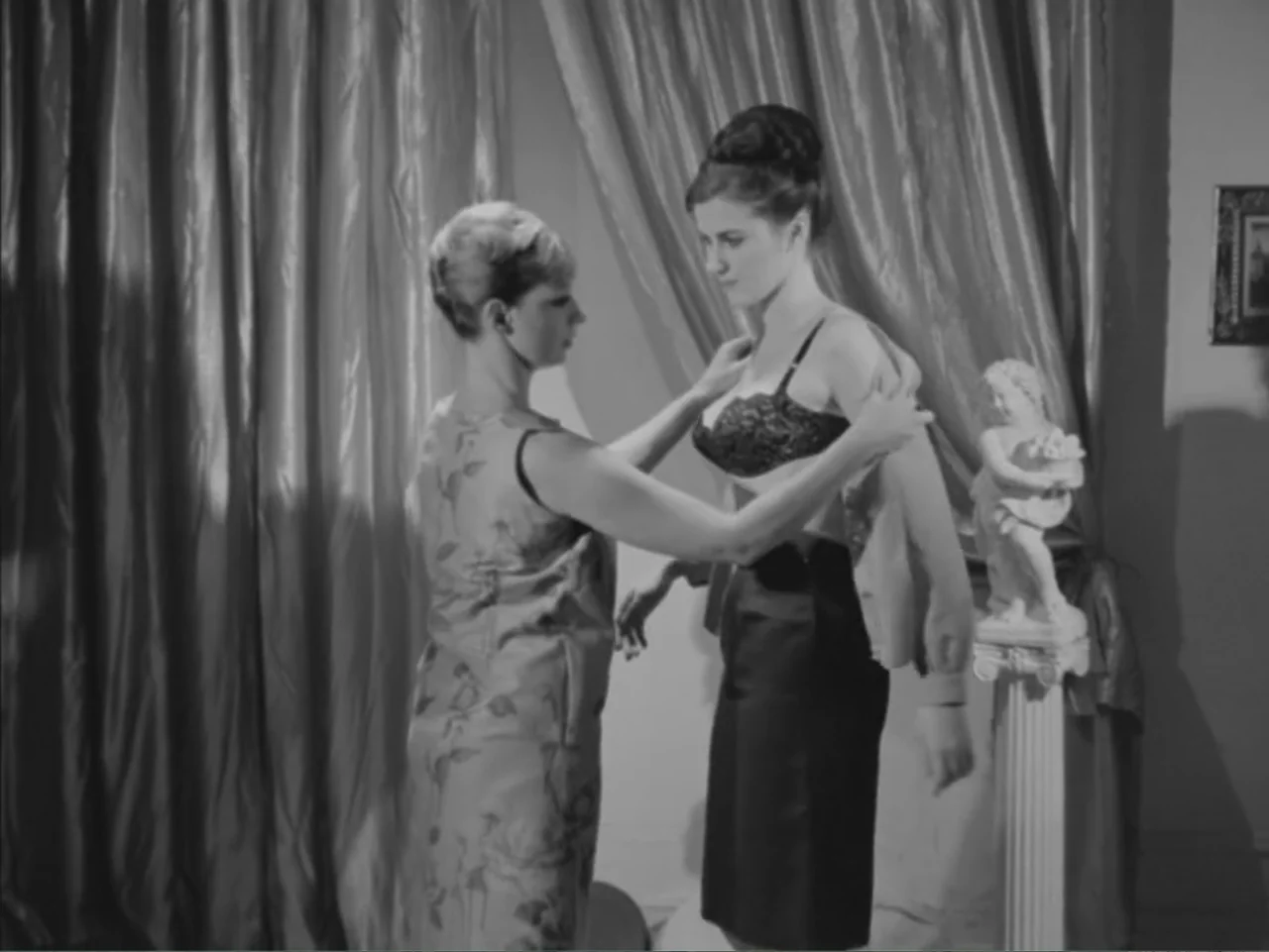 Chained Girls (1965) movie still: fullbody — A room features draped curtains and a small classical statue on a pedestal. One woman helps another…