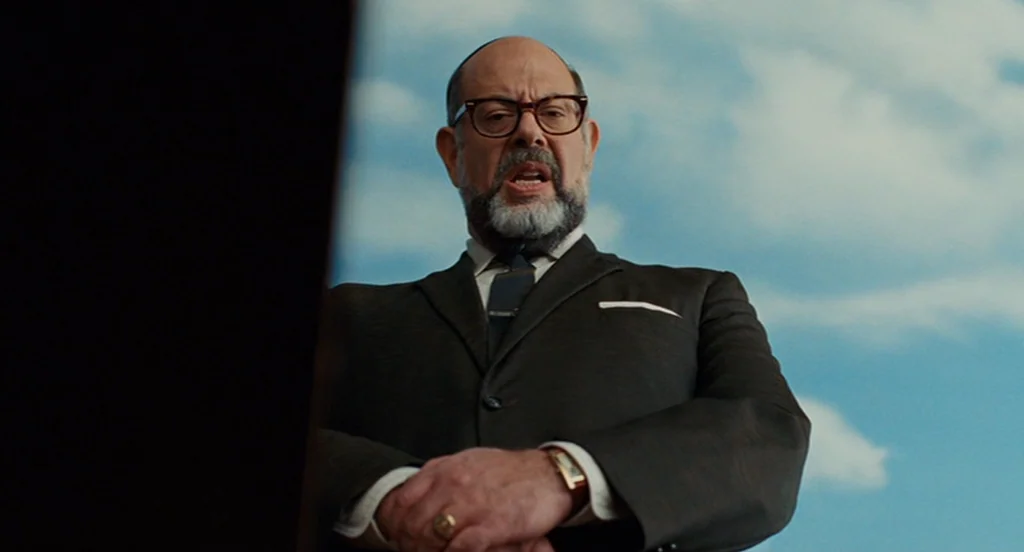 A Serious Man (2009) movie still: medium — An older man stands against a bright blue sky filled with scattered white clouds. The man in a formal…