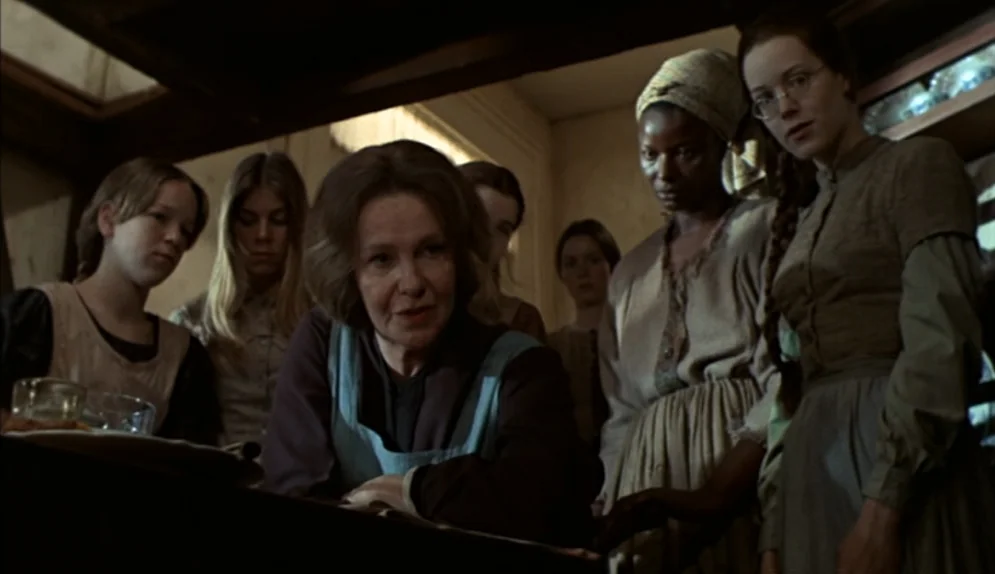 The Beguiled (1971) movie still: medium — A group of women in 19th-century attire gather around a wooden surface indoors. An older woman in an apron…