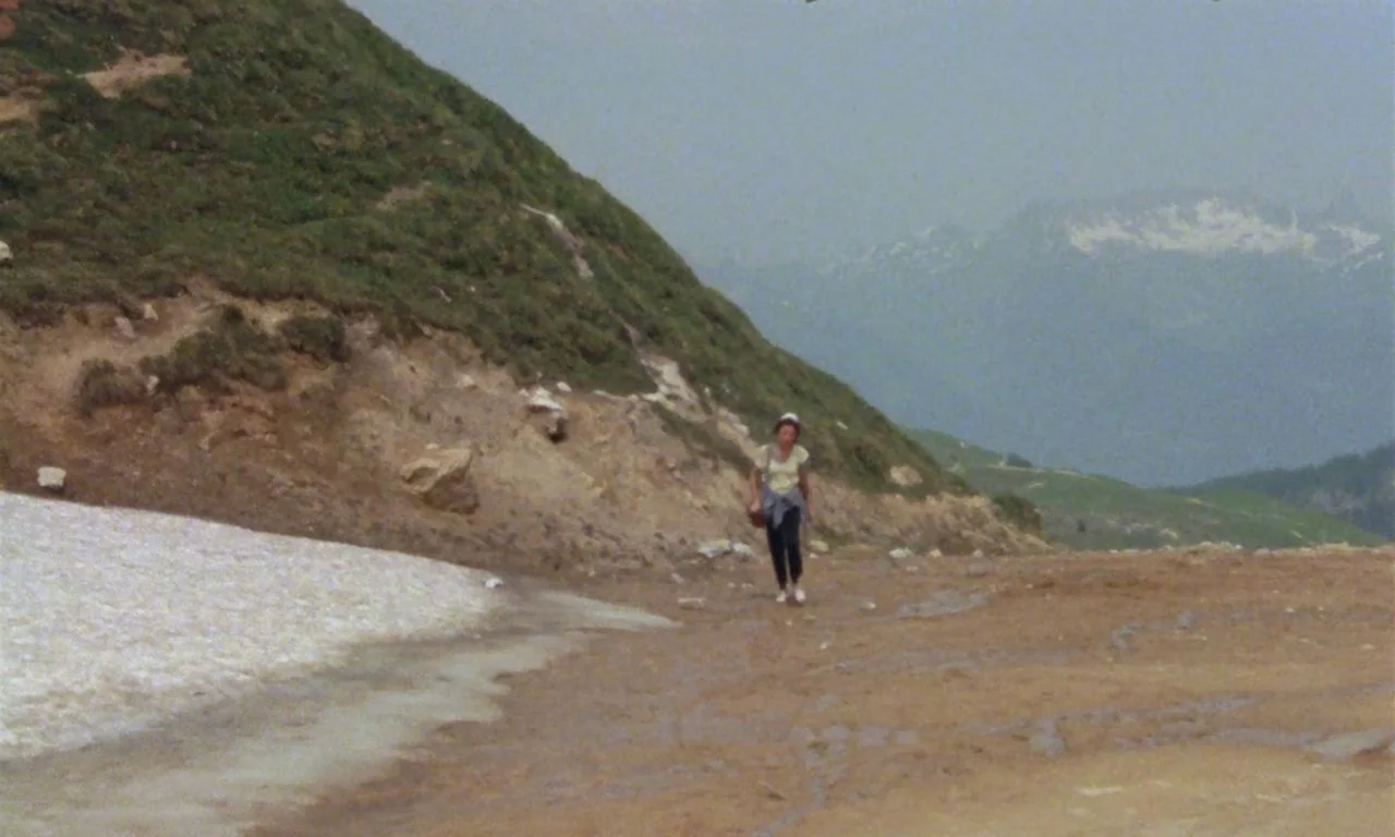 The Green Ray (1986) movie still: wide — A high-altitude mountain landscape features a patch of snow and a worn dirt path. A woman walks slowly…