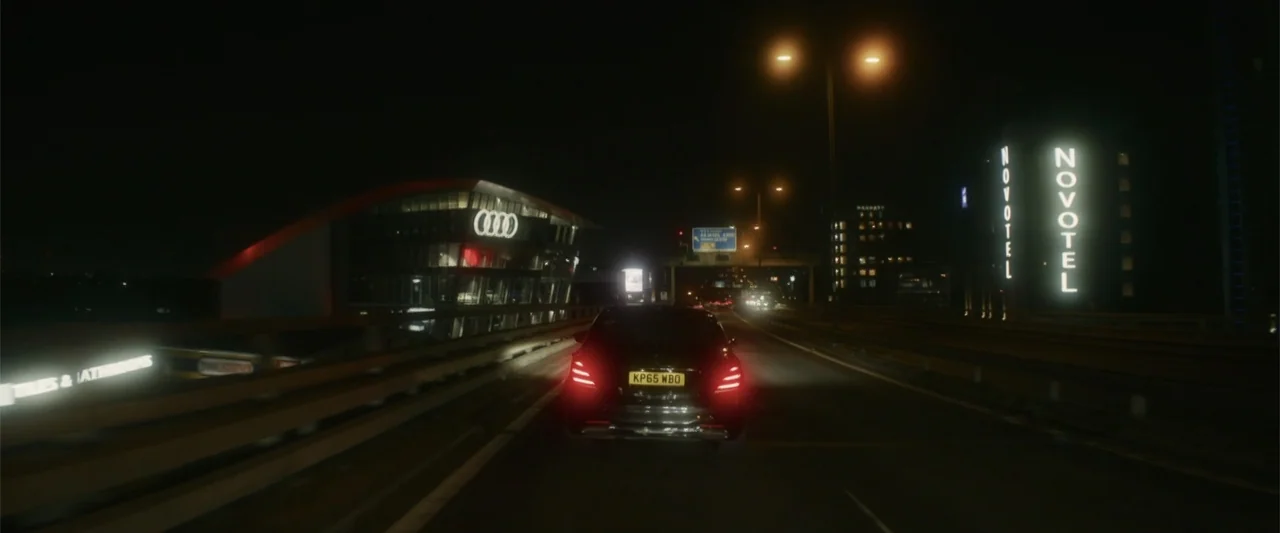 Black Bag (2025) movie still: medium — A night view from behind a car driving on a multi-lane urban highway. Glowing corporate building signs and…