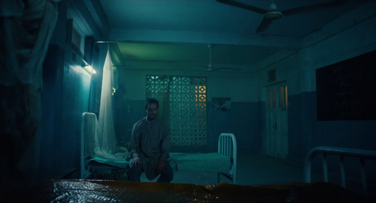 Submergence movie still: wide — A sparsely furnished ward features metal beds and a block-pattern window at night. A man sits motionless on the…