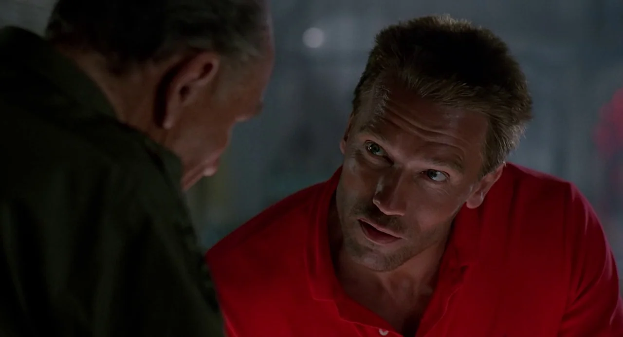 Predator (1987) movie still: medium — An interior space with dim, low-key lighting and blurred concrete surfaces in background. A man in a red shirt…