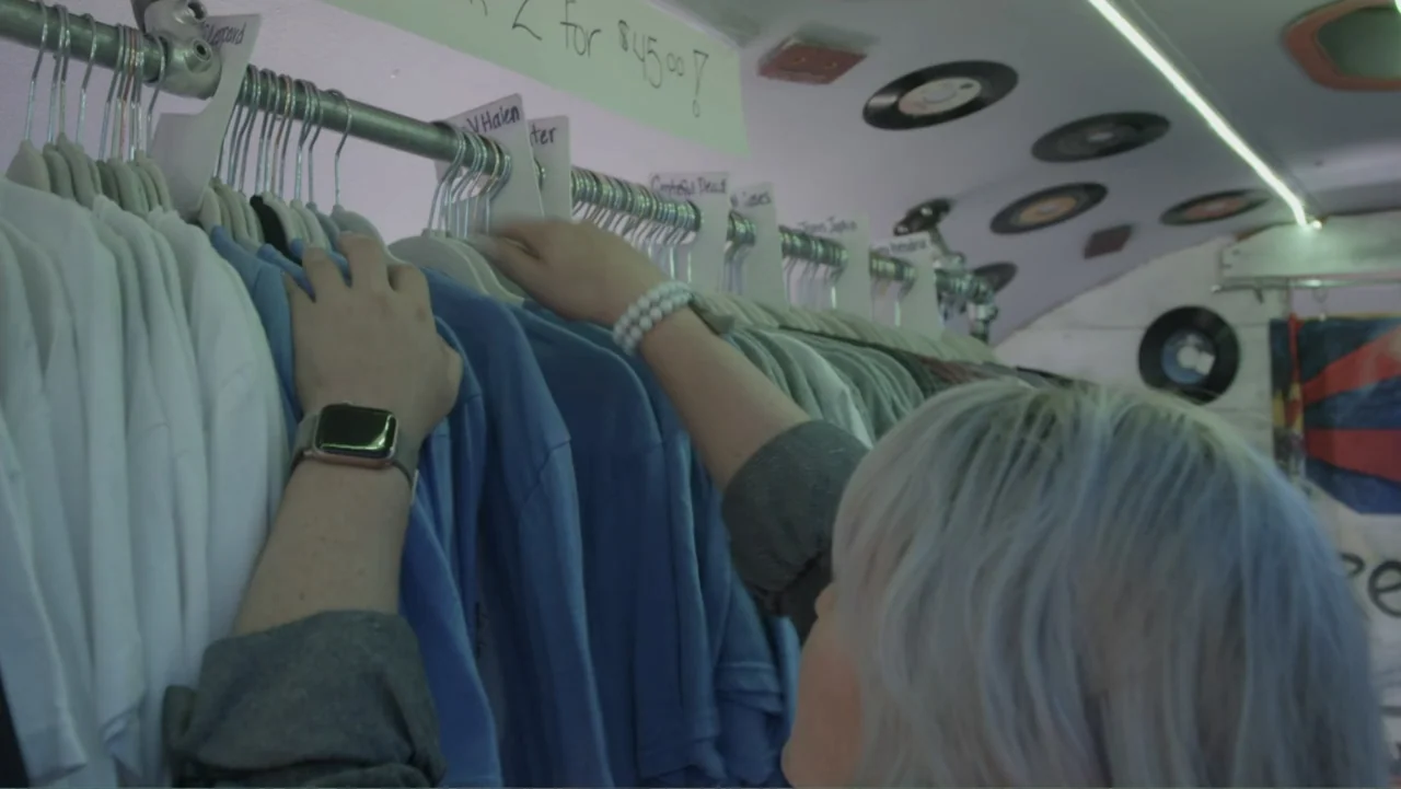 The Rise and Fall of Lularoe (2021) movie still: medium — A retail rack holds organized shirts with labels for bands above them. A person with light…