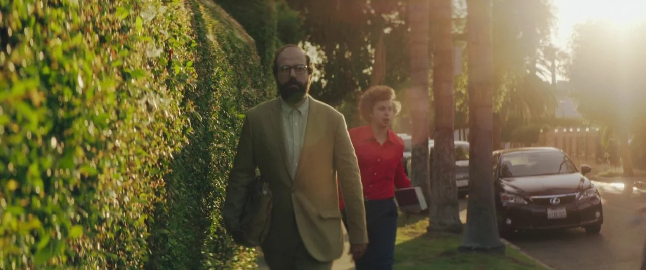 Lemon (2017) movie still: fullbody — A suburban sidewalk is lined with a thick manicured hedge and tall palm trees. Two men walk side-by-side along…