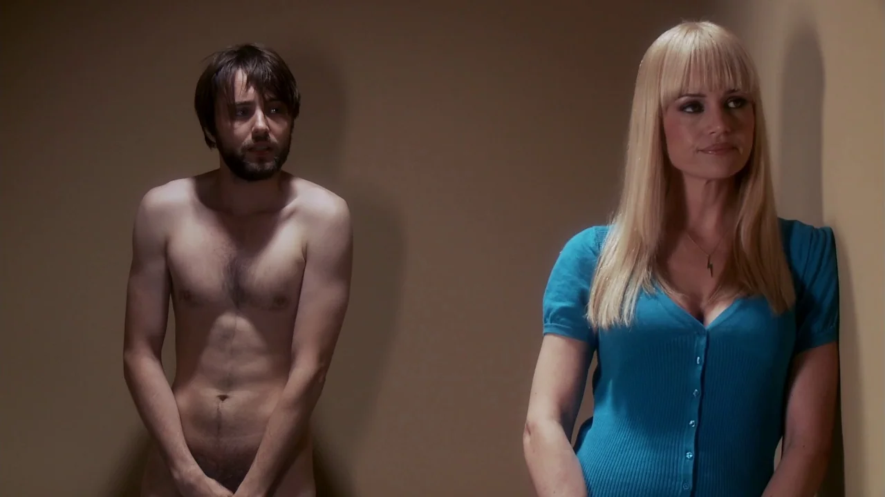 Electra Luxx (2010) movie still: medium — Two individuals stand in a minimalist room with featureless tan walls. A man stands without clothing to…