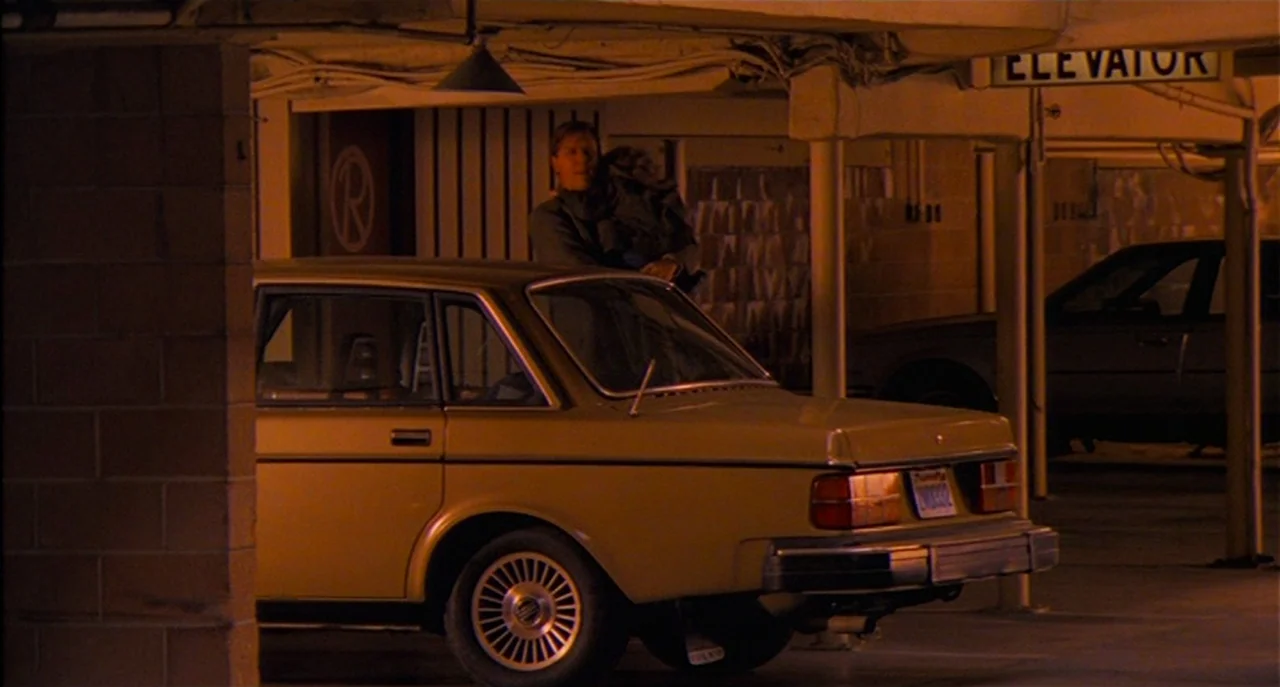 Raising Cain movie still: wide — A dimly lit parking structure features concrete pillars and an elevator entrance sign. A man stands behind a yellow…