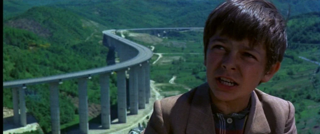Don’t Torture A Duckling (1972) movie still: medium — A green mountainous landscape features a large concrete highway viaduct curving through the…
