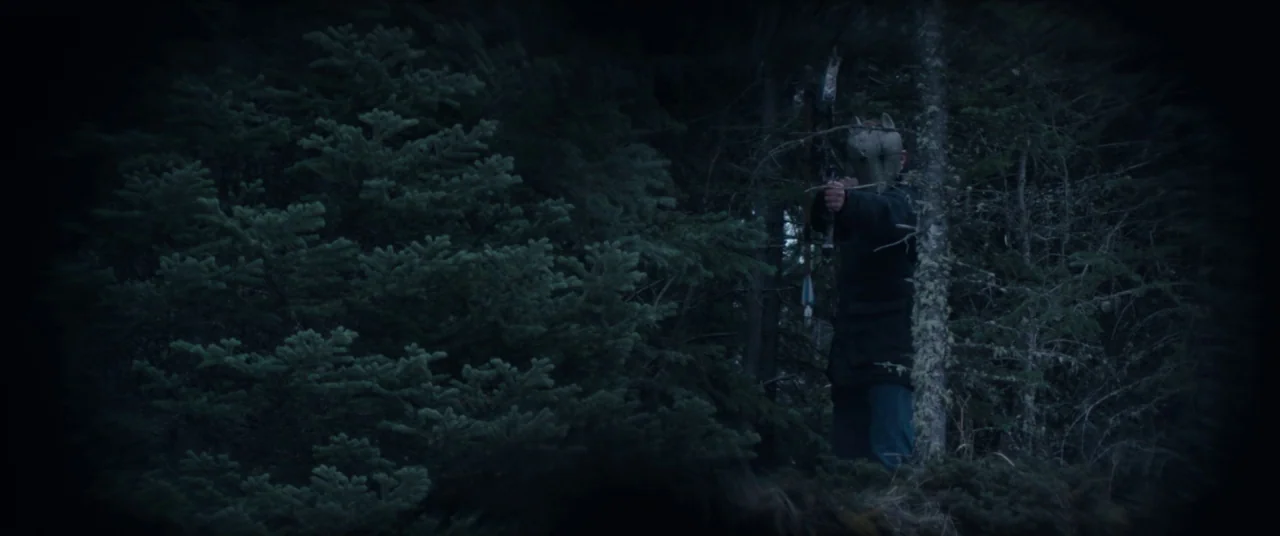 Hold The Dark (2018) movie still: medium — A dense, dark patch of evergreen woodland at night with heavy vignette-style lighting. A lone figure…
