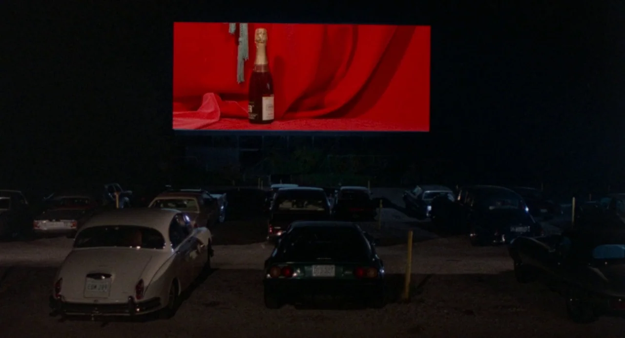 Polyester (1981) movie still: wide — A drive-in movie theater at night shows a bright red image of a champagne bottle. Several vintage and…
