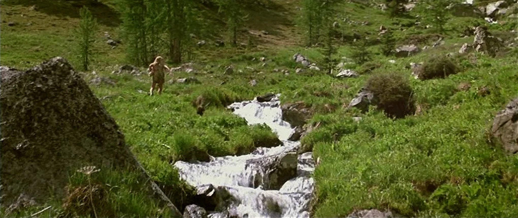 Clan of the Cave Bear (1986) movie still: wide — A high altitude grassy mountain slope features a small, rushing stream carving through rocks. A man…