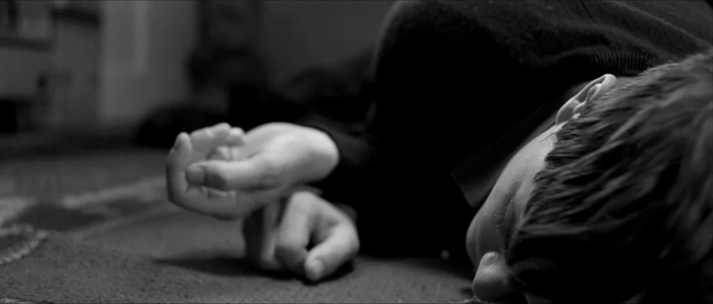 Control movie still: closeup — A man lies on a carpeted floor in a dark interior space. His head is turned to the side while his hands rest near his…