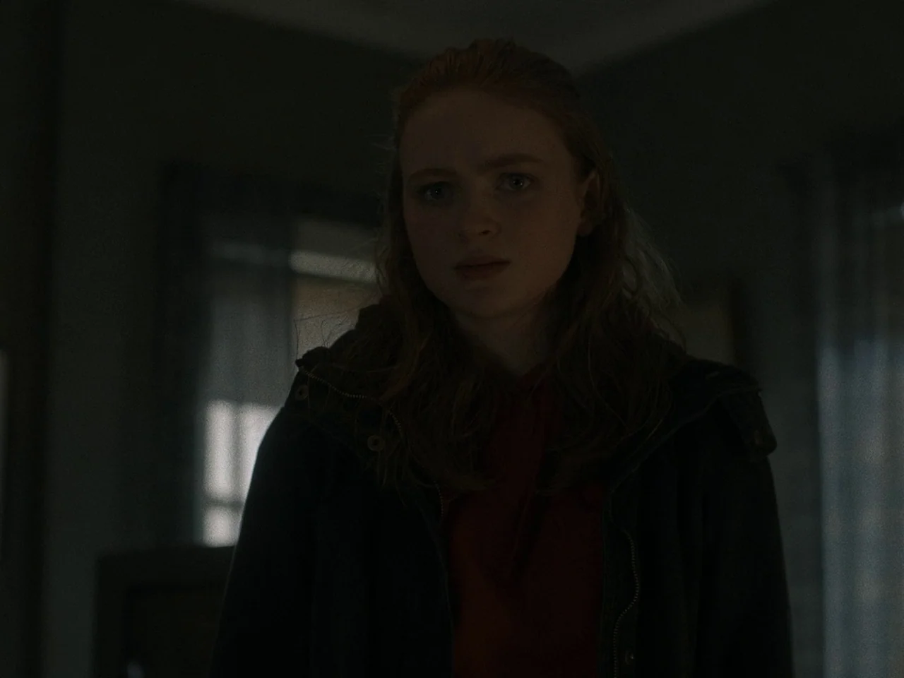 The Whale (2022) movie still: medium — A dimly lit interior space contains a young woman with shoulder-length wavy red hair. She wears a dark jacket…