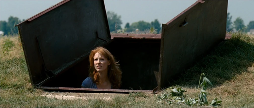 Take Shelter (2011) movie still: medium — A metal storm shelter hatch is open in a grassy field. A woman looks out from within the dark underground…