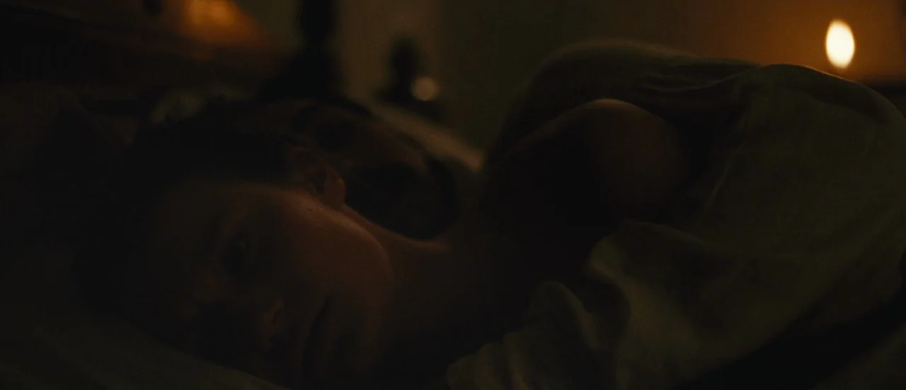 Madame Bovary movie still: closeup — A dark bedroom with two people lying in bed under white sheets. The woman faces the camera while the man rests…