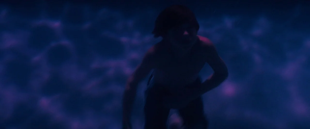 Honey Boy (2019) movie still: medium — A person swims beneath the surface of a pool at night. The dark water is illuminated by vibrant purple light…