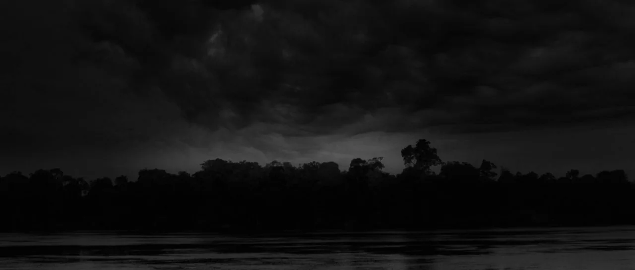 Embrace Of The Serpent (2015) movie still: establishing — A dark river flows beneath a thick, heavy layer of storm clouds. Dense silhouettes of…