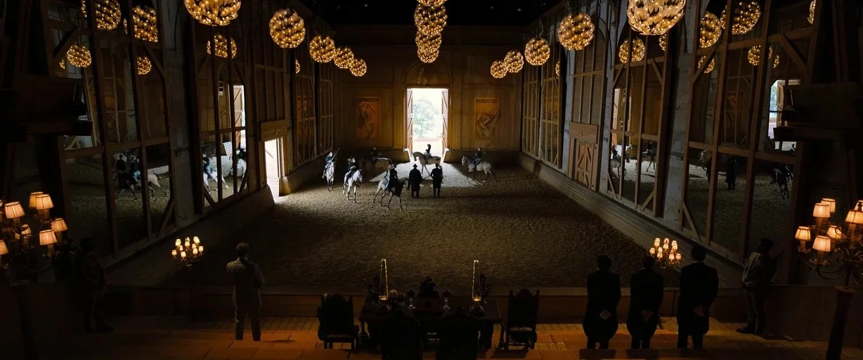 John Wick: Chapter 4 (2023) movie still: wide — A spacious indoor riding hall features sand flooring and large walls lined with mirrors. Men in…