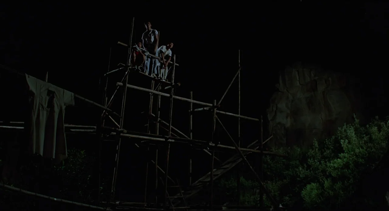 Typhoon Club (1985) movie still: wide — Three young men sit atop a tall bamboo scaffolding structure at night. A large rock formation is visible to…