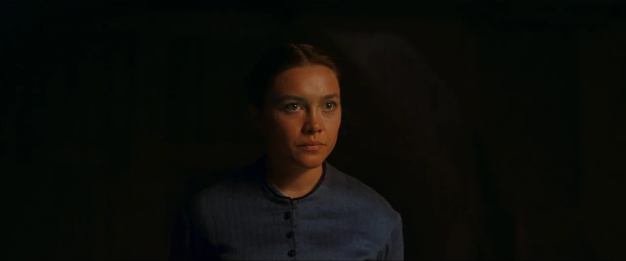 The Wonder (2022) movie still: medium — A woman stands in a dark, indistinct interior setting with minimal environmental detail. The figure is…