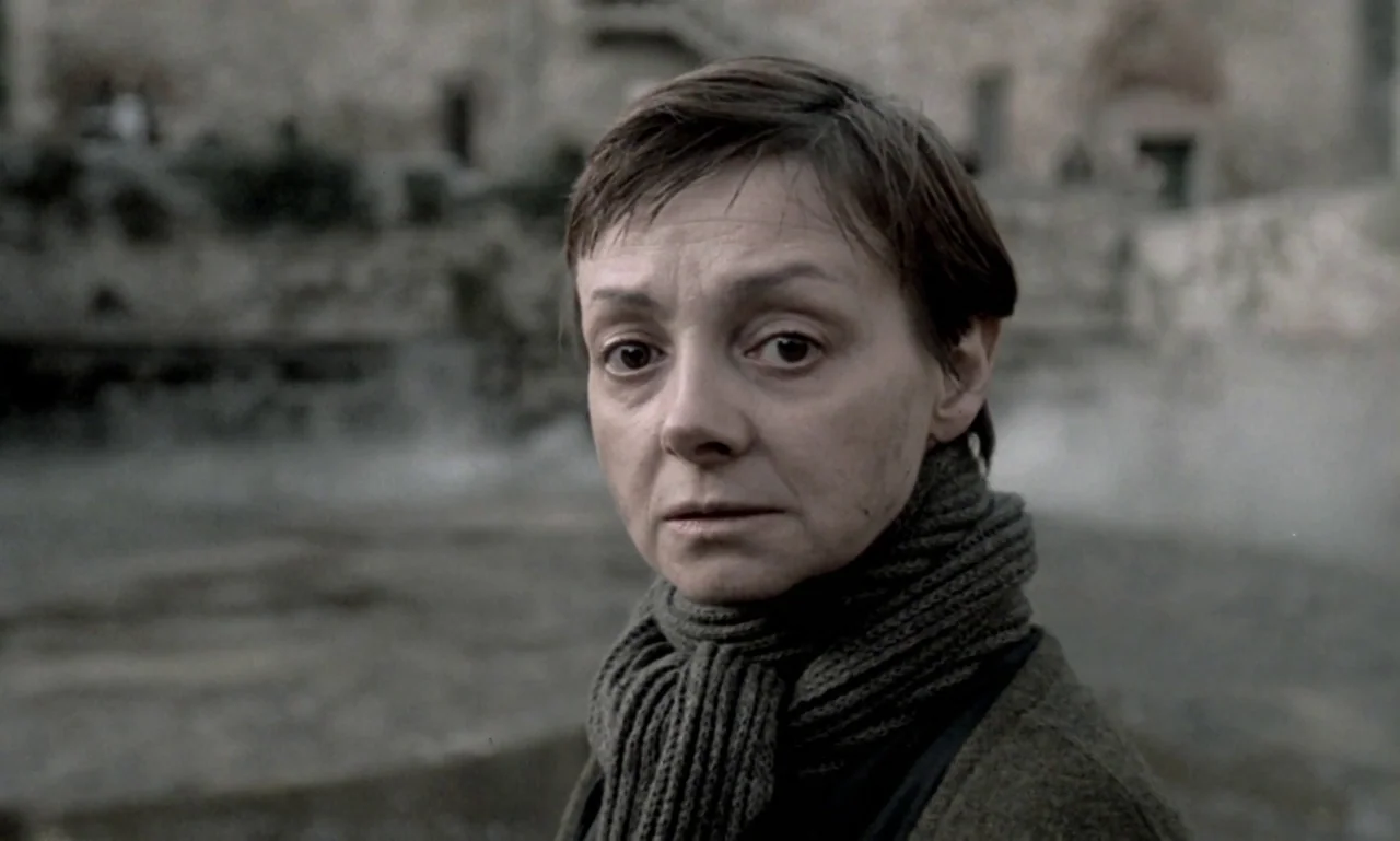 Nostalghia (1983) movie still: closeup — A stone courtyard provides an overcast, textured backdrop behind the subject. A woman looks forward against…