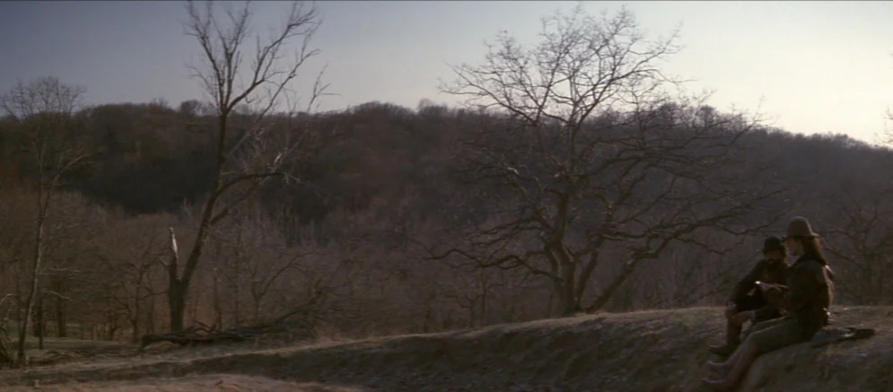 Ride With The Devil movie still: wide — A desolate hillside overlooks a dense forest of leafless trees under a pale sky. Two men sit on a dirt ridge…