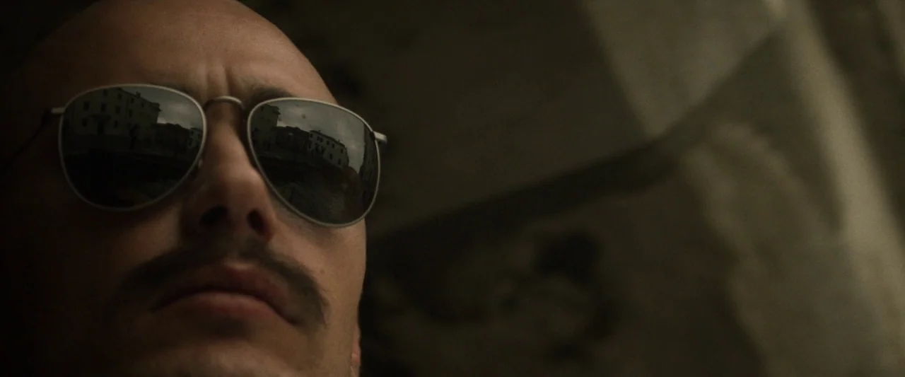 Zeroville (2019) movie still: closeup — A close-up shot focuses on a man wearing metallic-framed sunglasses against a shadowed background. The…
