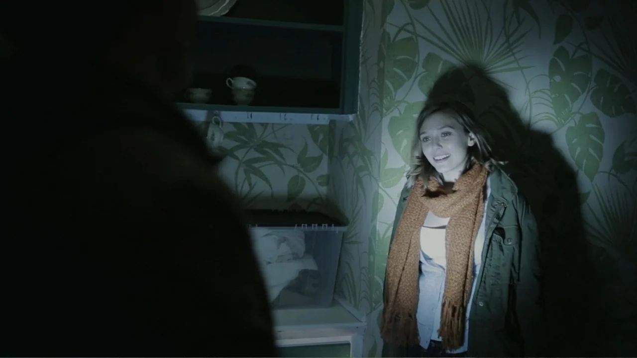 Silent House (2011) movie still: medium — A small corner of a room features green leaf-patterned wallpaper and open shelves. A woman illuminated by…