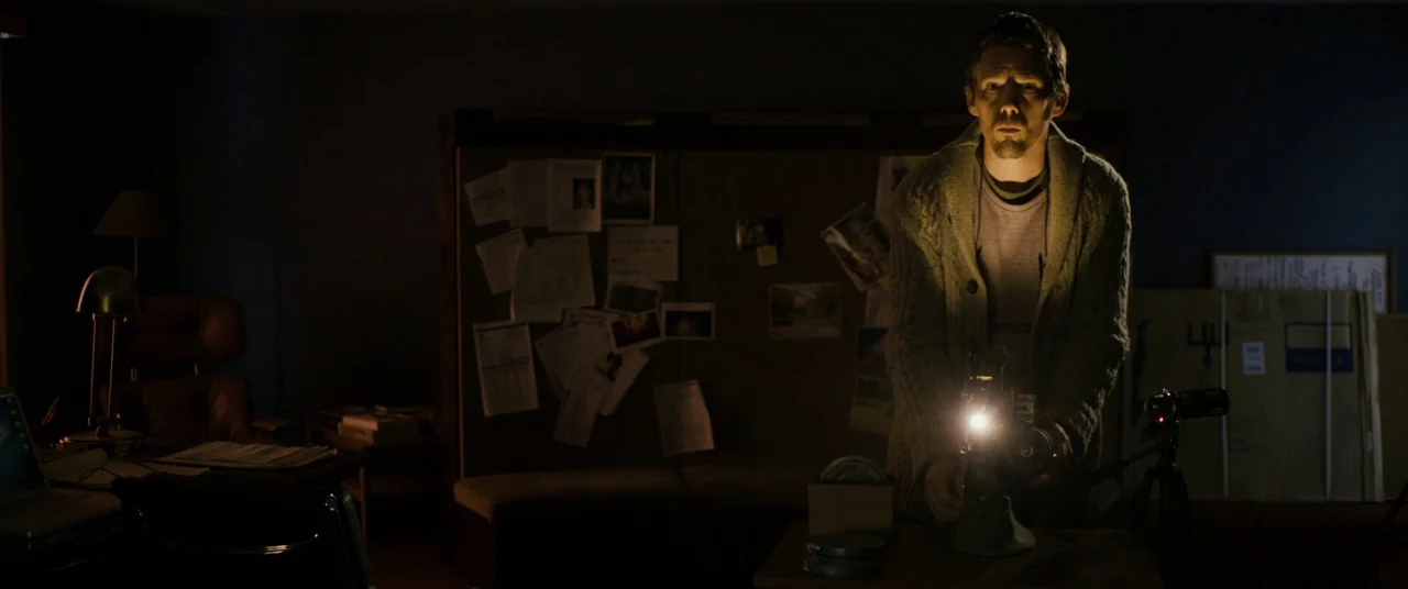 Sinister (2012) movie still: medium — A dark, cluttered room contains a pin-up board covered in papers and photographs. A middle-aged man stands at…