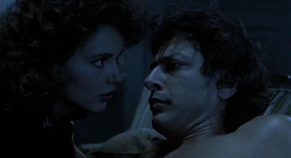 The Fly (1986) movie still: medium — A dark, dimly lit bedroom serves as the backdrop for this conversation. A woman and a man face each other…
