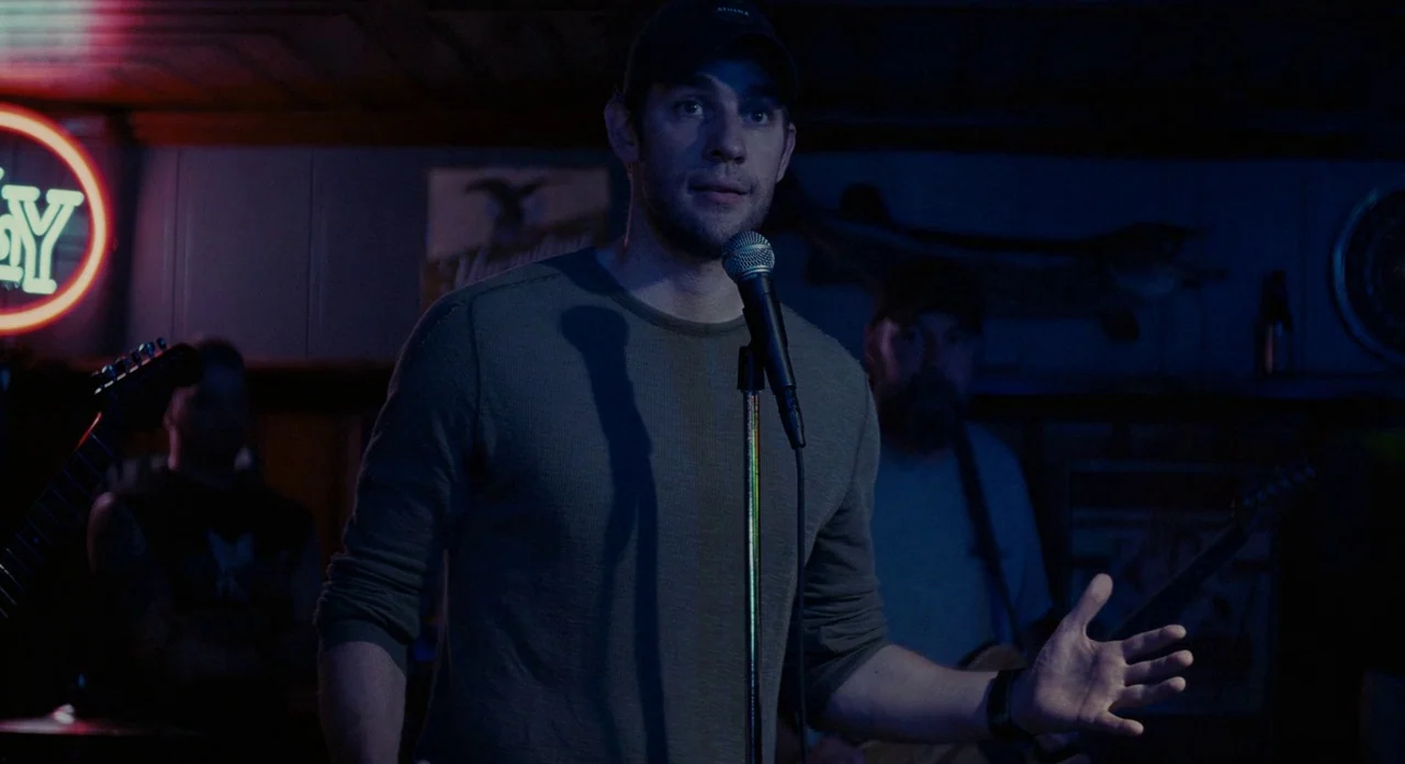 Promised Land (2012) movie still: medium — A dimly lit interior space contains a musician standing before a microphone stand. He addresses an unseen…