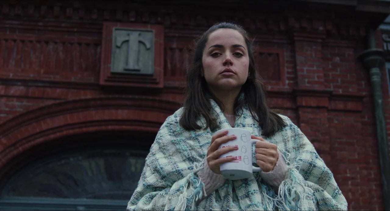 Knives Out (2019) movie still: medium — A woman stands in front of a dark red brick wall with a stone insignia. She wears a thick shawl and holds a…