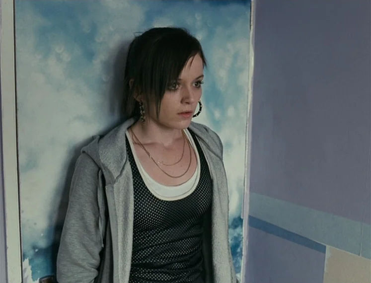 Fish Tank (2009) movie still: medium — A young woman stands against a door covered in a sky-patterned poster in a room. She faces slightly away from…