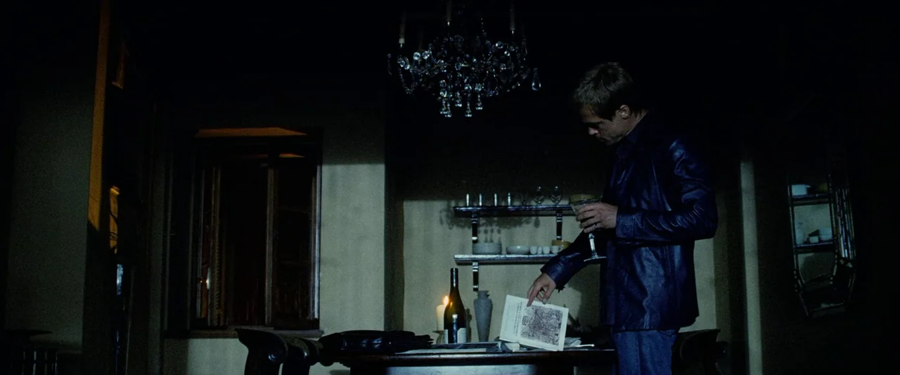 Ocean’s Twelve (2004) movie still: medium — A dimly lit room features a dark wooden table with an open book and wine. A man stands beside the table,…