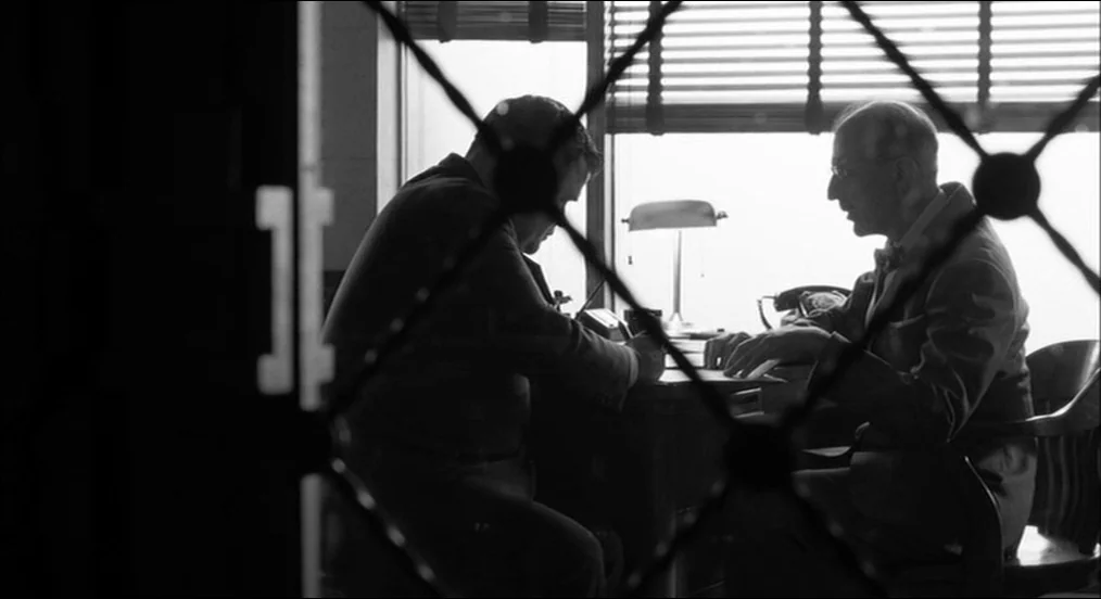 The Man Who Wasn’t There (2001) movie still: medium — A dark, cramped office interior viewed through a black metal security gate. Two men sit at a…