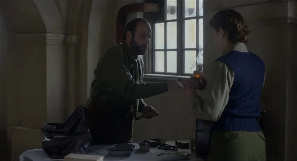 The Innocents (2016) movie still: medium — A stone-walled room features a table covered with medical instruments and supplies. A bearded man hands a…